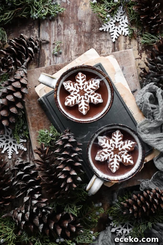 Two mugs of hot chocolate topped with snowflake marshmallow on a wood table surrounded by pinecones.