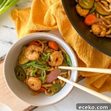 A white bowl of Shrimp Yaki Udon sits on a rustic wooden cutting board with chopsticks with a yellow linen and green onions.