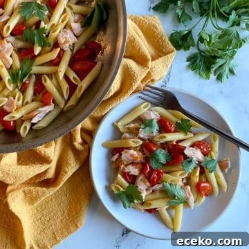A white plate of penne pasta with burst cherry tomatoes and flaked salmon topped with parsley sits on a white marble countertop.