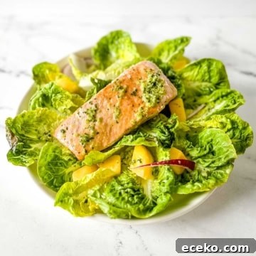A fillet of salmon sits on a bed of little gem lettuce, mango, red onion, and jalapeno vinaigrette on a white marble counter.