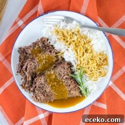 a white plate half full of roast beef and half full of rice. Roast beef and rice are both covered with gravy. There is a fork on the plate next to the rice. The plate is on an orange napkin with white stripes.