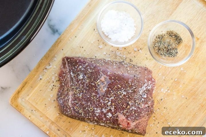 An uncooked beef roast sprinkled with salt and pepper on a wooden cutting board next to a slow cooker. There is a small glass bowl of salt and a small glass bowl of pepper on the cutting board next to the roast.