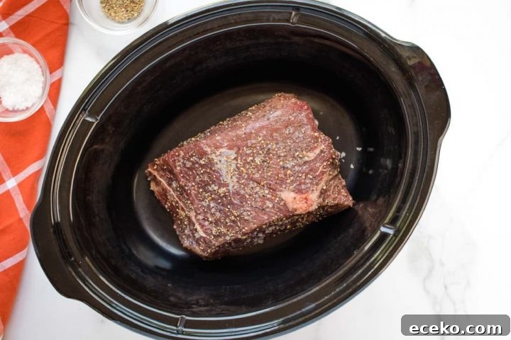Uncooked beef roast in a slow cooker on a white marble counter. On the left side of the photo is an orange napkin with white stripes. On the napkin is a small glass bowl with salt. Next to the napkin is small glass bowl with pepper.