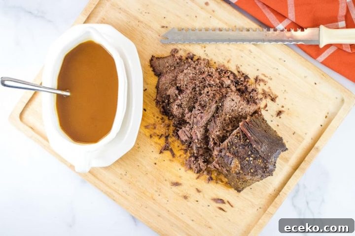 White gravy bowl on a white platter. The bowl has brown gravy and a spoon. The platter is sitting on a wooden cutting board next to a beef roast that has been half-sliced with the serrated knife at the top of the photo. There is an orange napkin with white stripes in the top right corner of the photo.
