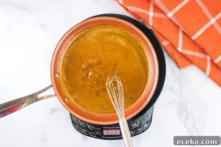 Overhead shot of a small orange skillet of brown gravy over a warming unit on a white marble counter.There is a whisk in the skillet, and an orange napkin with white stripes on the counter at the top right of the picture.
