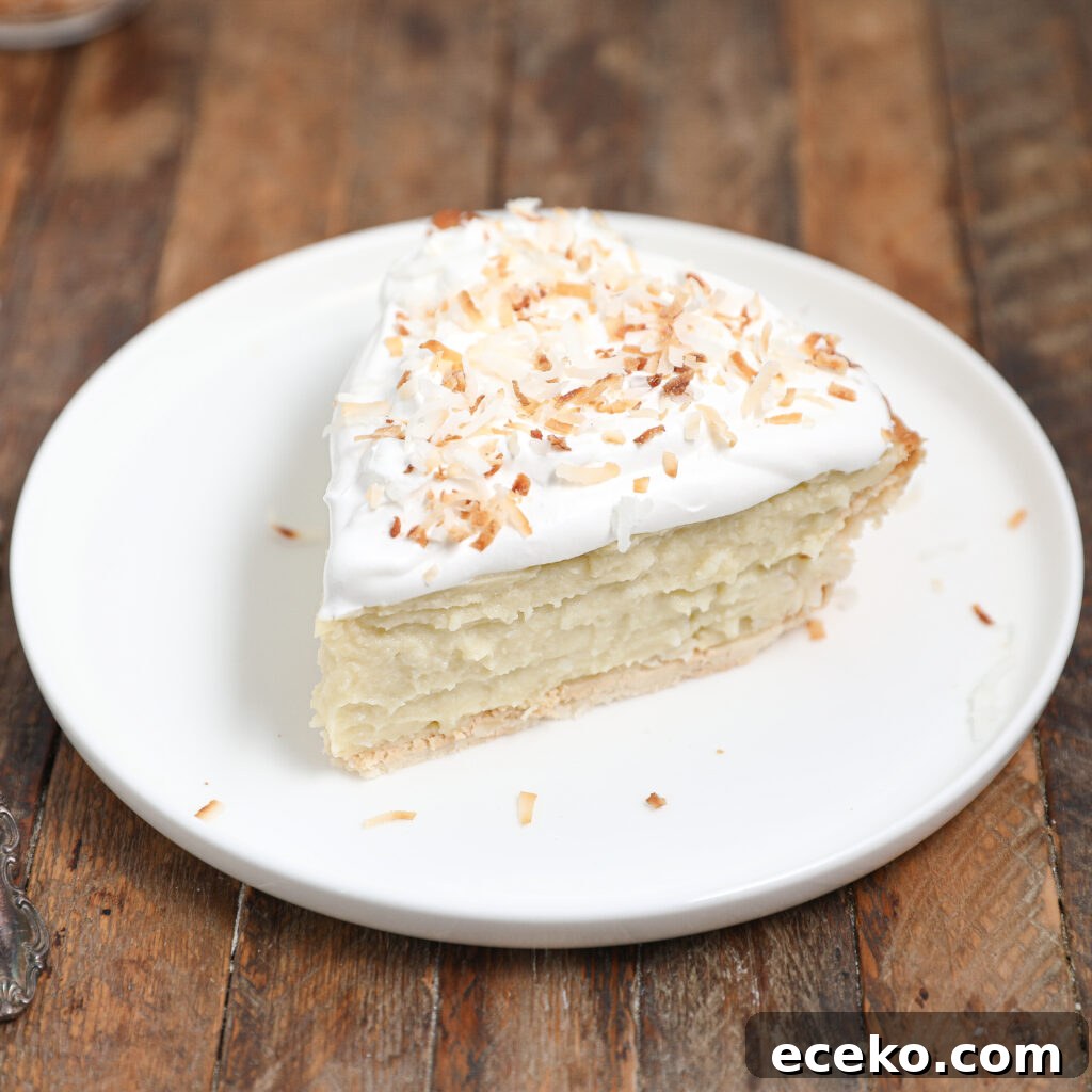 A slice of Coconut Cream Pie with whipped topping and toasted coconut flakes sits on a white plate atop a rustic wooden table.