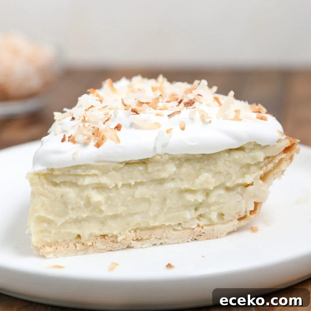 A close-up of a Coconut Cream Pie slice with a flaky crust, creamy coconut filling, whipped topping, and toasted coconut flakes on a white plate.