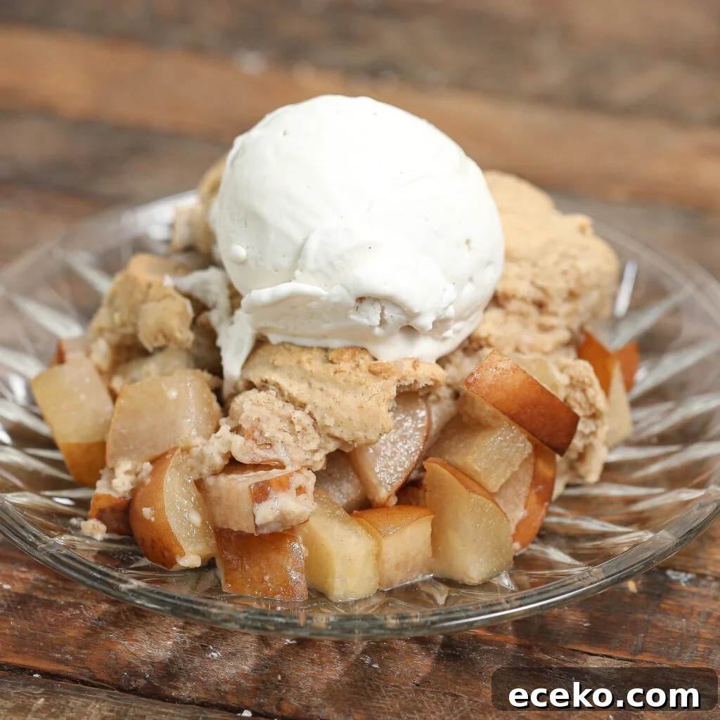Warm Pear Cobbler with Cinnamon and Brown Sugar 2 A glass plate with baked diced pears and a biscuit-like topping, this Pear Cobbler is finished with a scoop of vanilla ice cream and sits on a rustic wooden surface.