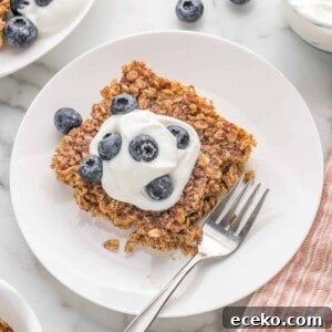 Square of baked oatmeal on a white plate, topped with yogurt and fresh blueberries.