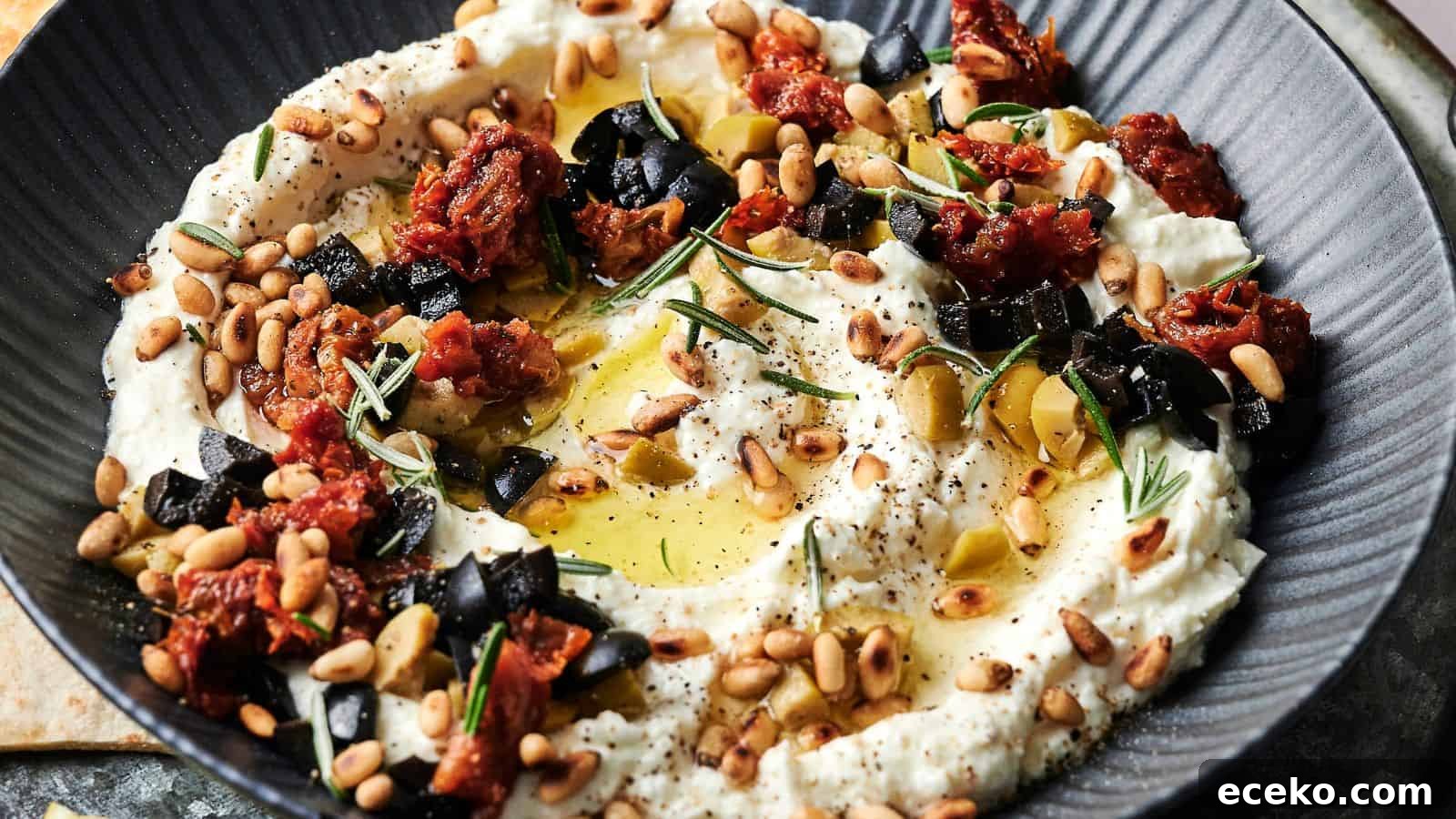 A bowl of creamy dip garnished with chopped black olives, sun-dried tomatoes, pine nuts, rosemary, olive oil, and black pepper.
