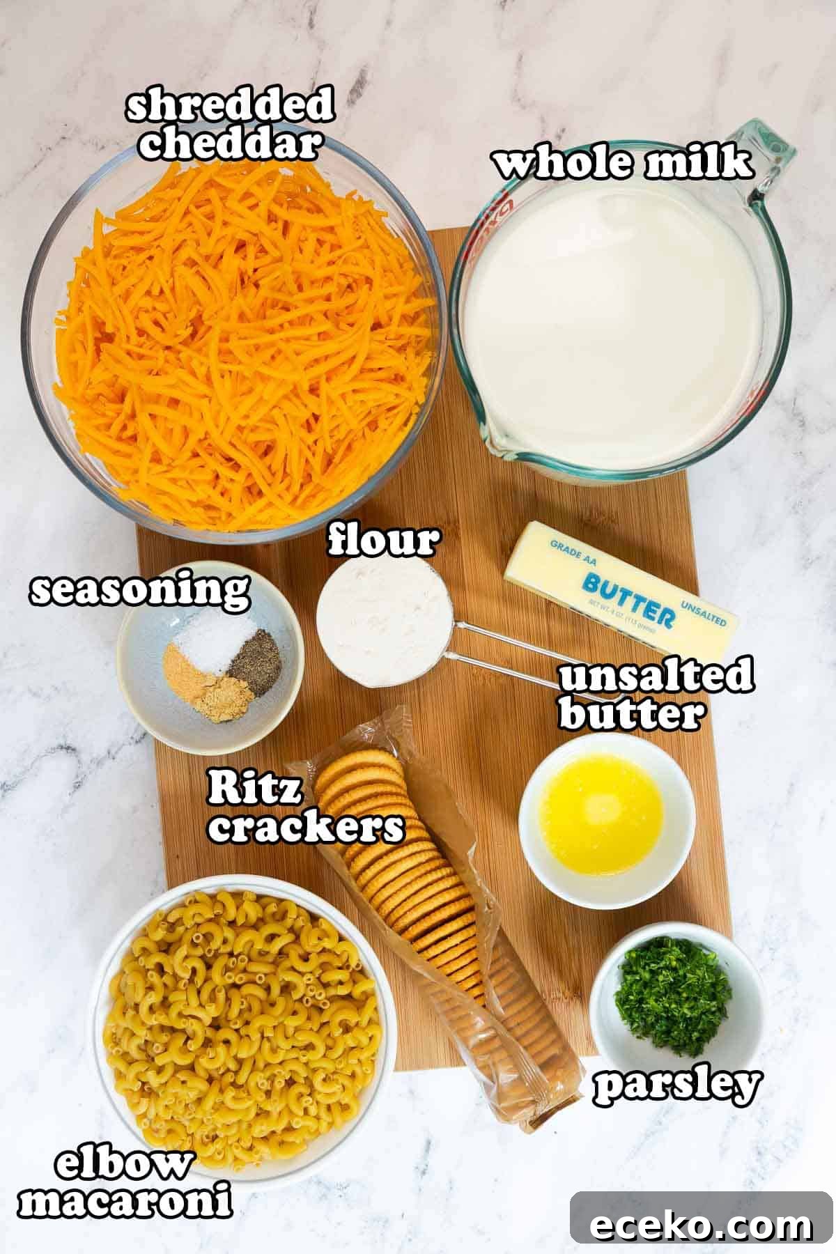 Ingredients for old fashioned baked mac and cheese with Ritz crackers displayed on a wooden board, including shredded cheddar, whole milk, unsalted butter, flour, elbow macaroni, Ritz crackers, seasoning, and parsley.