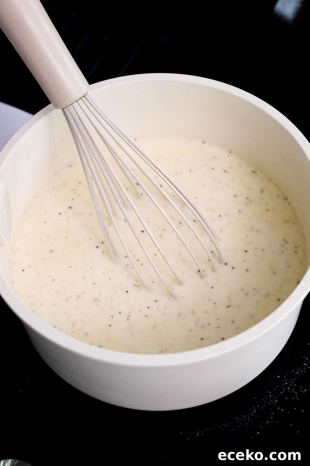 Flour and seasoning whisked into milk to create a béchamel-style base in a white saucepan during the early stages of making mac and cheese sauce.