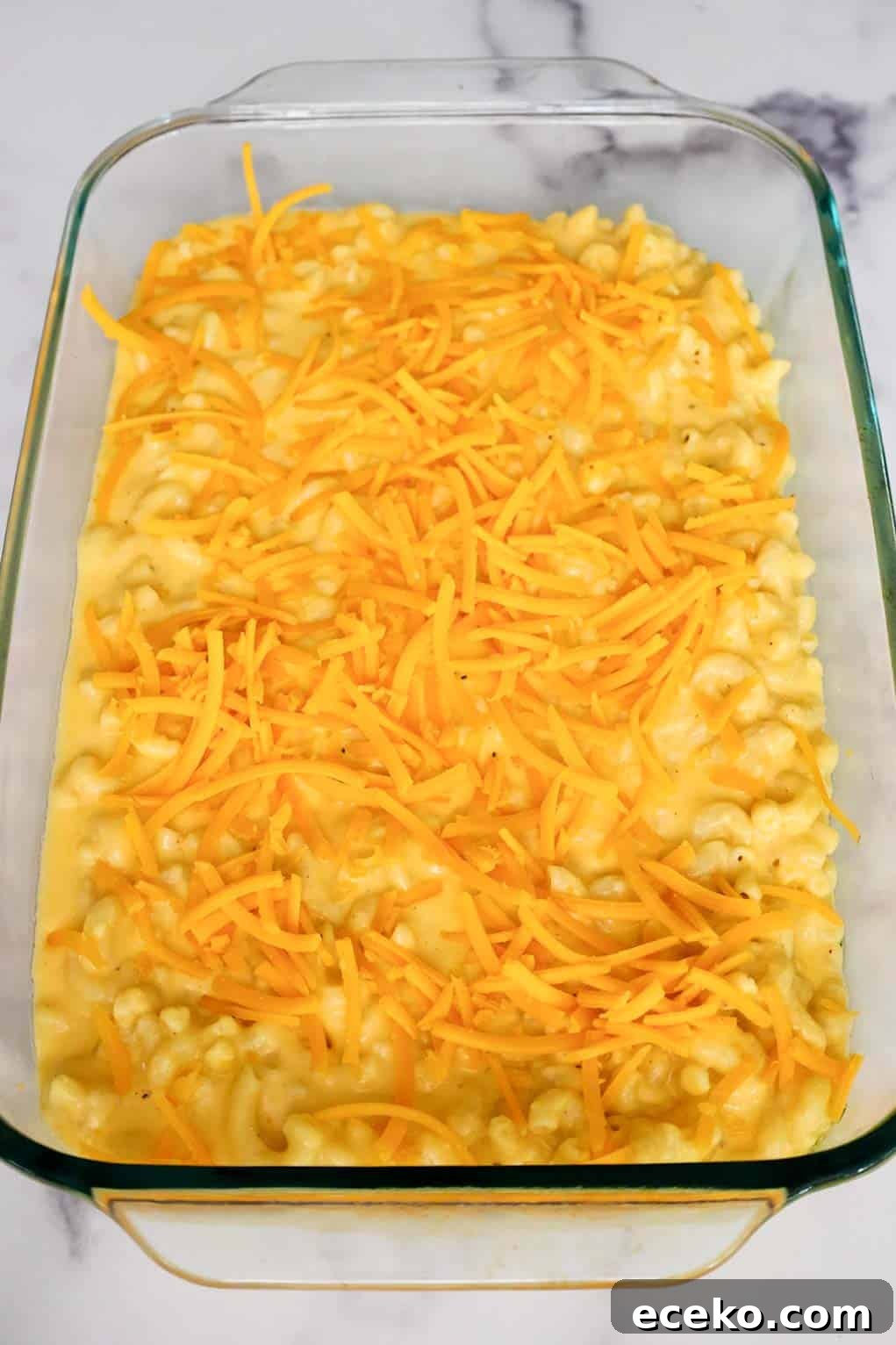Unbaked old fashioned mac and cheese in a glass baking dish topped with shredded cheddar cheese, ready for the Ritz cracker topping.