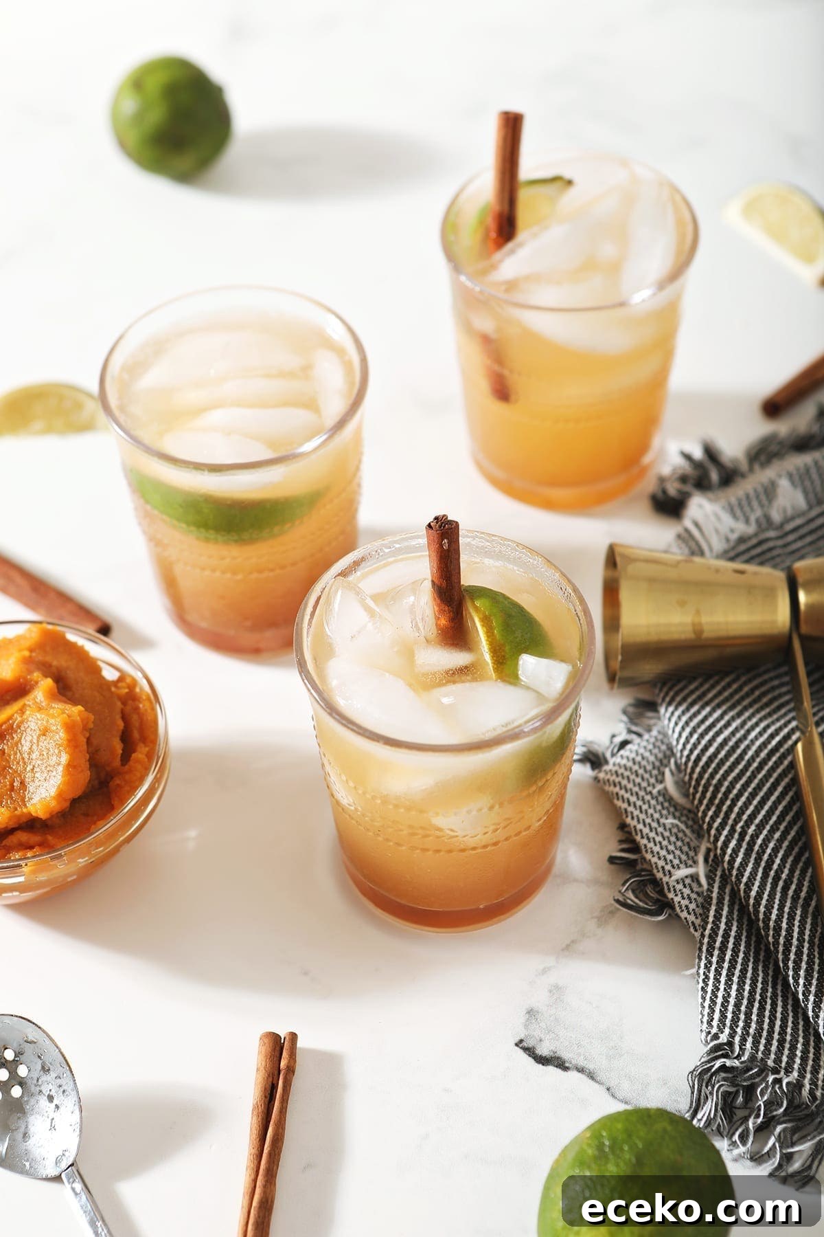 Three Pumpkin Mules, garnished with cinnamon sticks and lime wedges, surrounded by limes, pumpkin and a jigger on marble