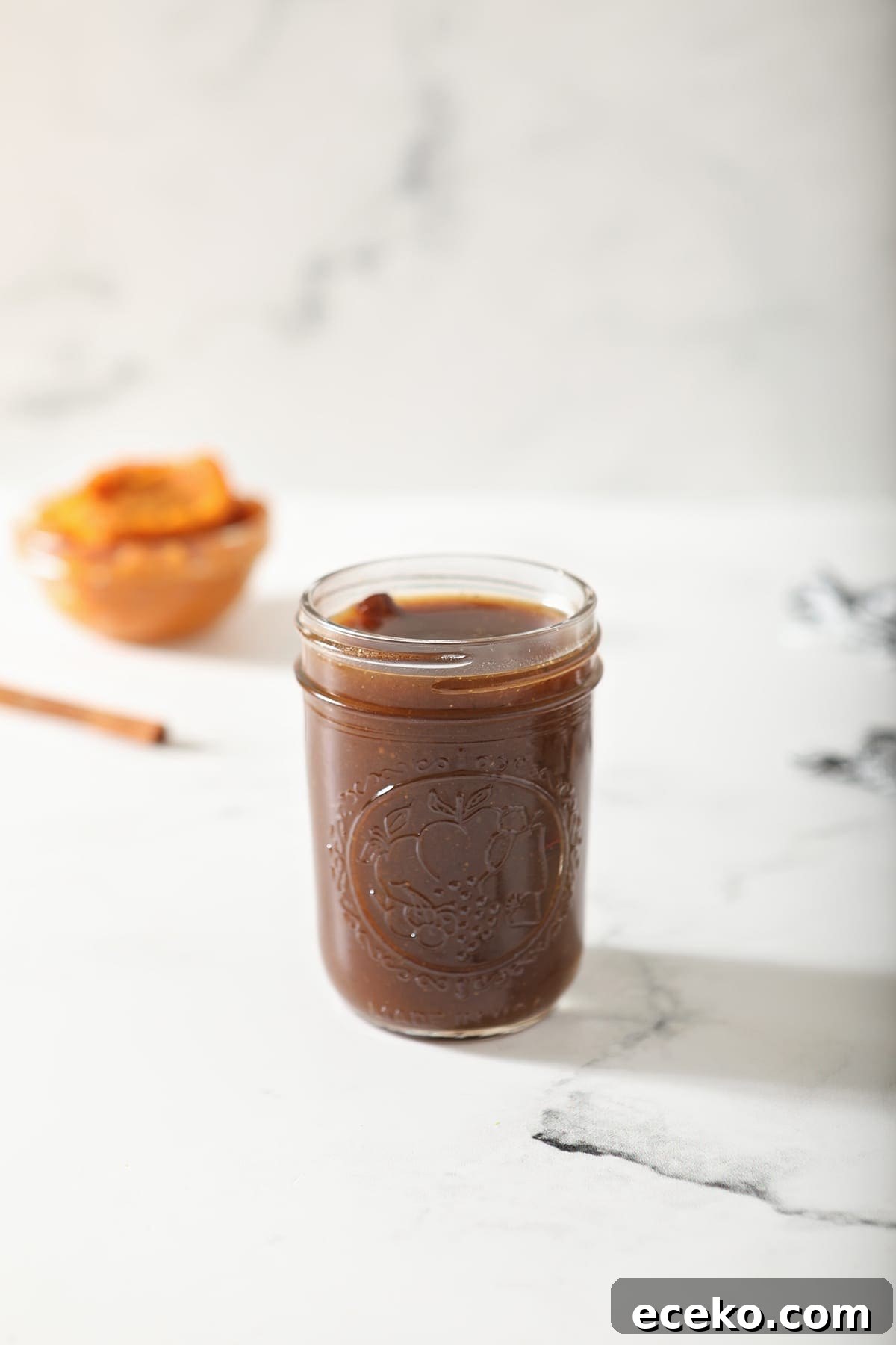 A jar of Pumpkin Spice Syrup sits on marble with a cinnamon stick and pumpkin puree behind it