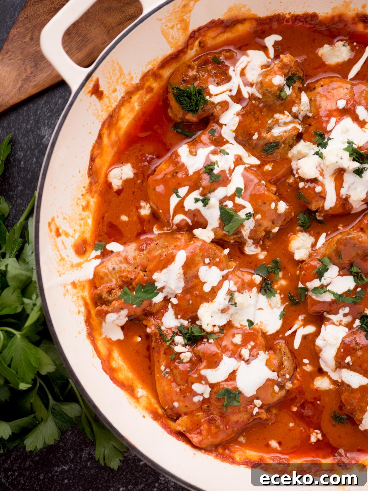buffalo chicken thighs in a skillet surrounded by fresh parsley, a serving utensil, and a kitchen towel