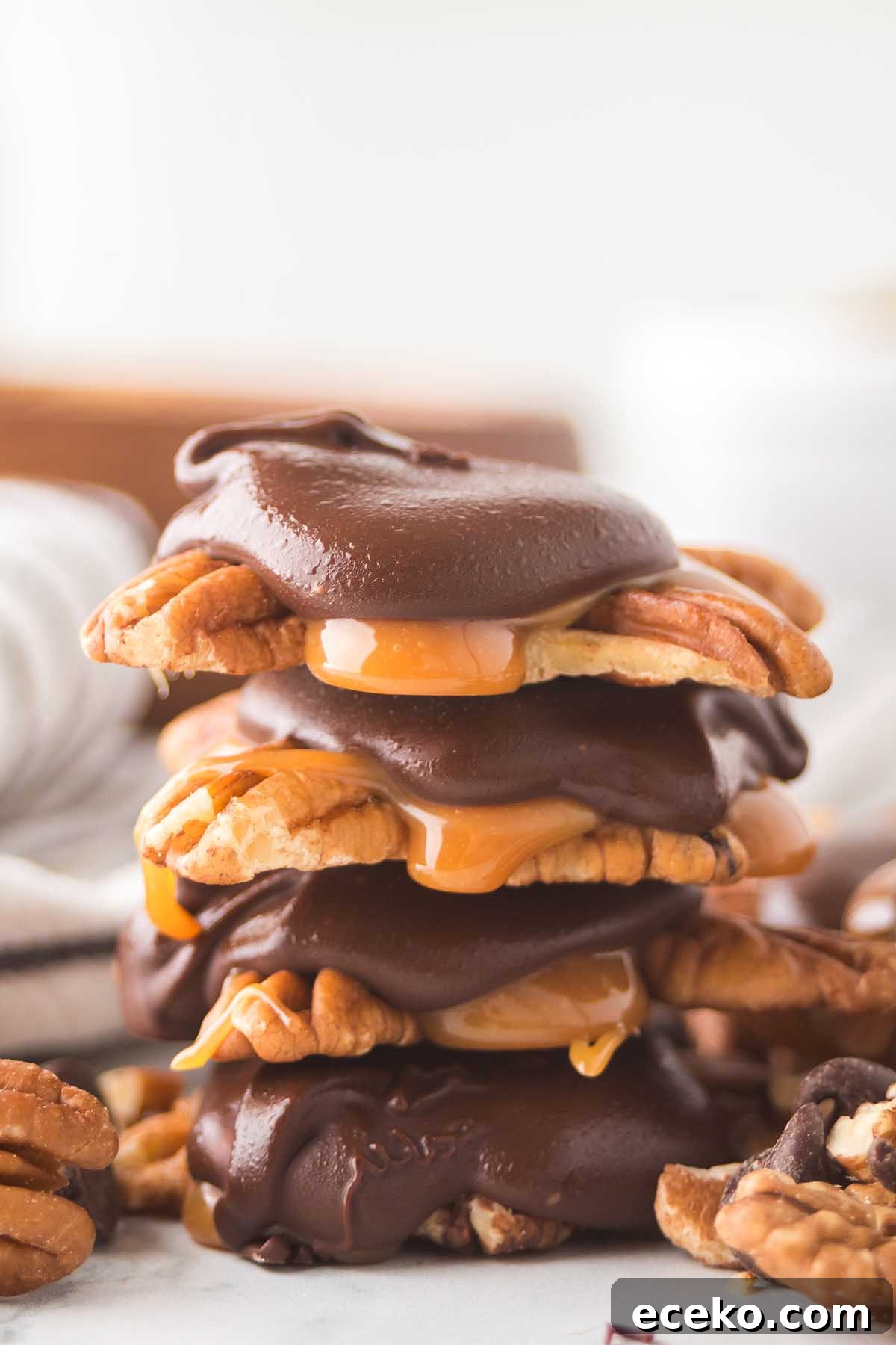 How to Make Classic Homemade Turtle Candies at Home 11 A stack of homemade turtles, rich with chocolate-covered caramel and nut clusters, rests on a white surface. Nearby, pecans and chocolate pieces add to the enticing display.