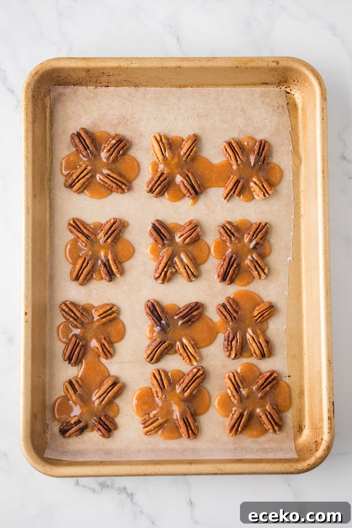 How to Make Classic Homemade Turtle Candies at Home 16 A baking tray with parchment paper holds twelve pecan pralines in flower shapes, reminiscent of homemade turtles.