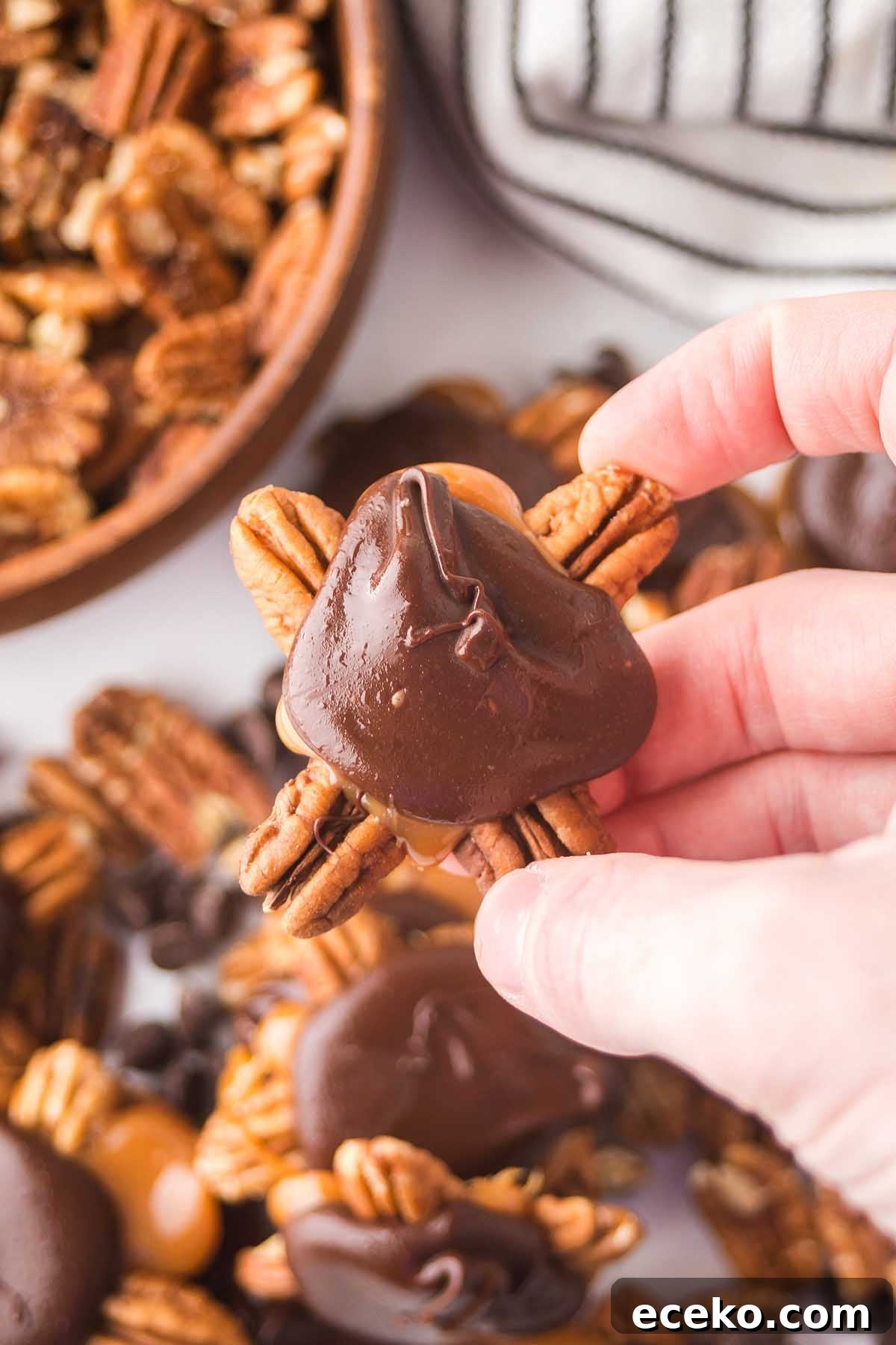 How to Make Classic Homemade Turtle Candies at Home 5 A hand holds a chocolate-covered pecan cluster with gooey caramel, reminiscent of homemade turtles, surrounded by more clusters and pecans in the background.