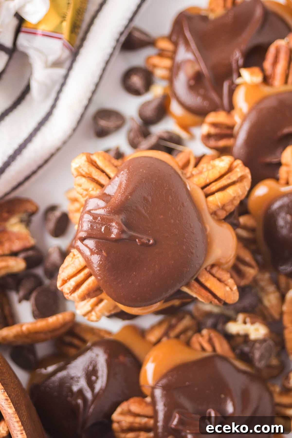 How to Make Classic Homemade Turtle Candies at Home 10 Close-up of homemade turtles featuring chocolate caramel pecan clusters arranged on a surface. A few chocolate chips and striped fabric can be seen in the background.