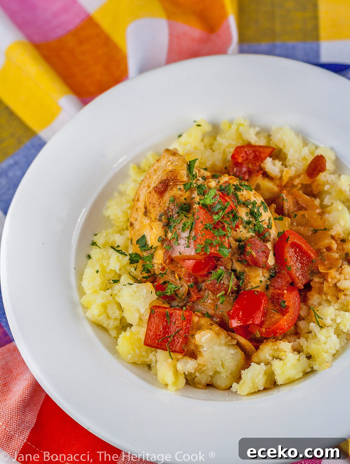A white bowl with smashed yellow Yukon Gold potatoes is topped with chicken braised in tomatoes, onions, mushrooms, and wine; Comforting Chicken Cacciatore with smashed Yukon Gold potatoes © 2023 Jane Bonacci, The Heritage Cook. 