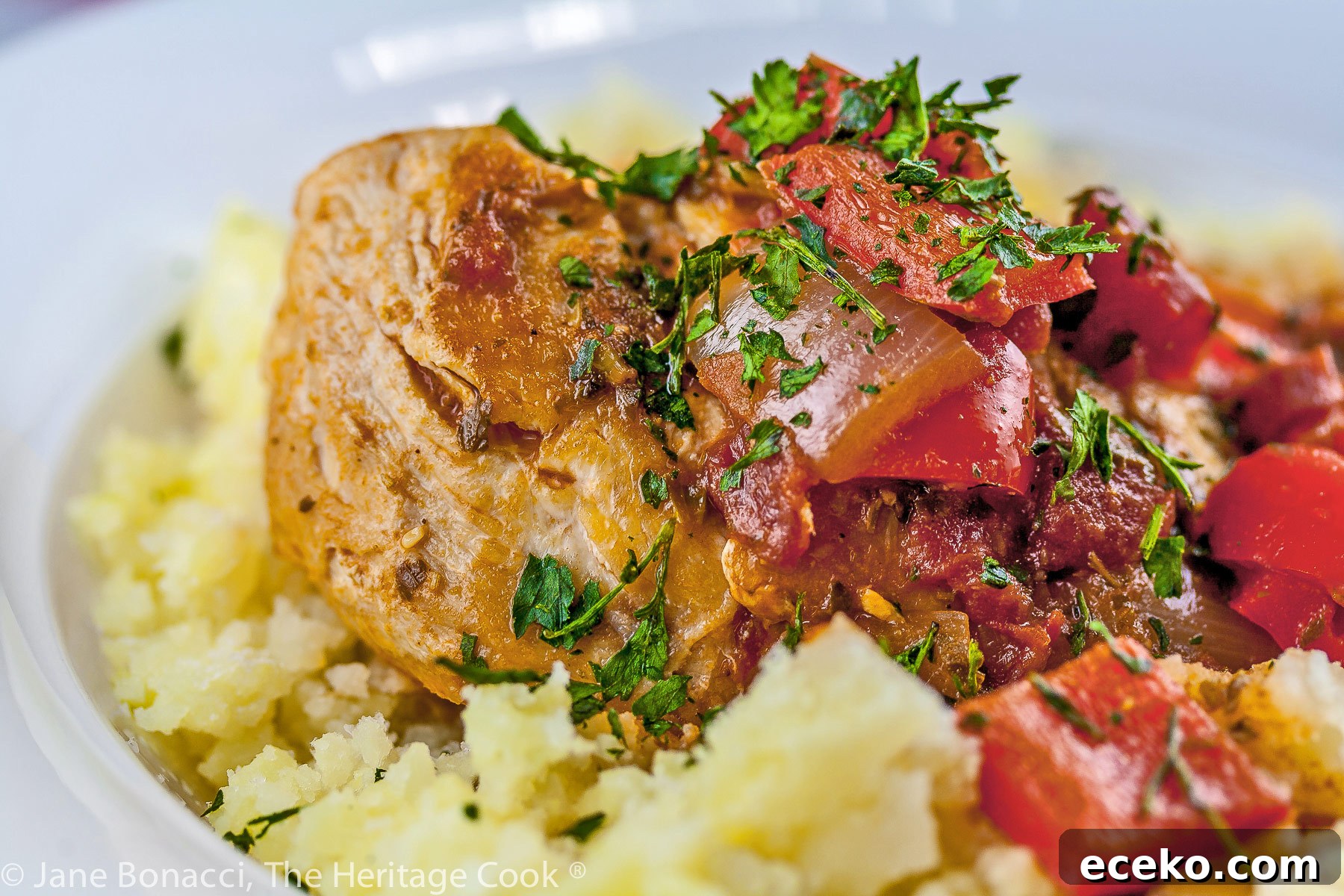 A white bowl with smashed yellow Yukon Gold potatoes is topped with chicken braised in tomatoes, onions, mushrooms, and wine; Comforting Chicken Cacciatore with smashed Yukon Gold potatoes © 2023 Jane Bonacci, The Heritage Cook. 