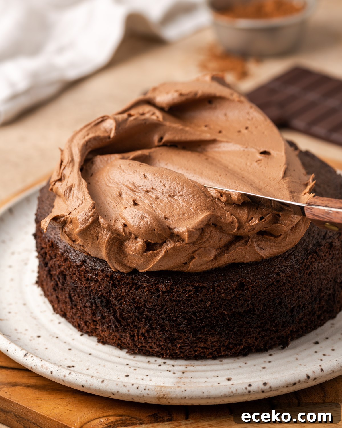 A large portion of chocolate frosting being spread on top of a baked chocolate cake.