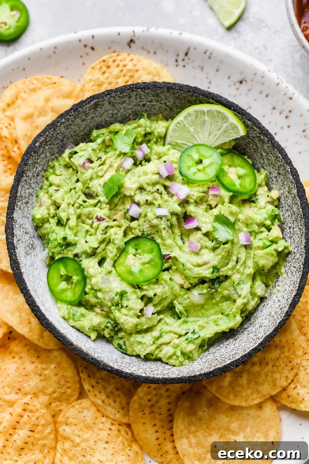 Overhead shot of a small bowl of bright green guacamole. Guacamole is topped with some finely diced onion, thinly sliced jalapeño, and cilantro leaves, surrounded by round yellow tortilla chips. 