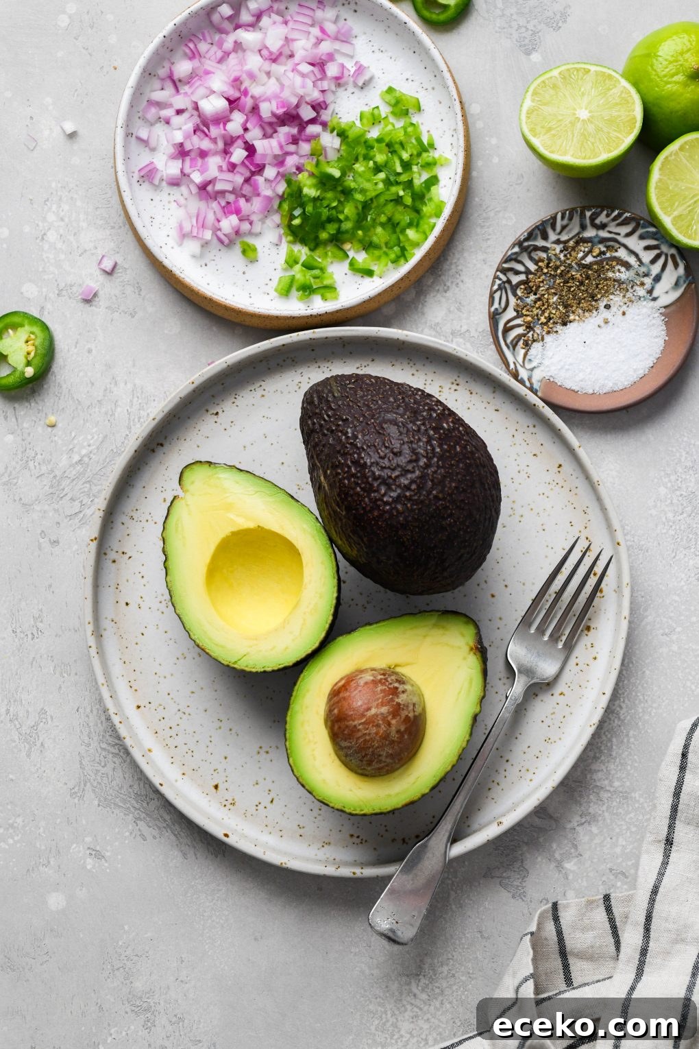 Image of ingredients used to make easy guacamole recipe. A plate with 2 avocados - one cut in half, a smaller plate with finely diced red onion, jalapeno, some cut limes, and a smaller plate with salt and pepper. 