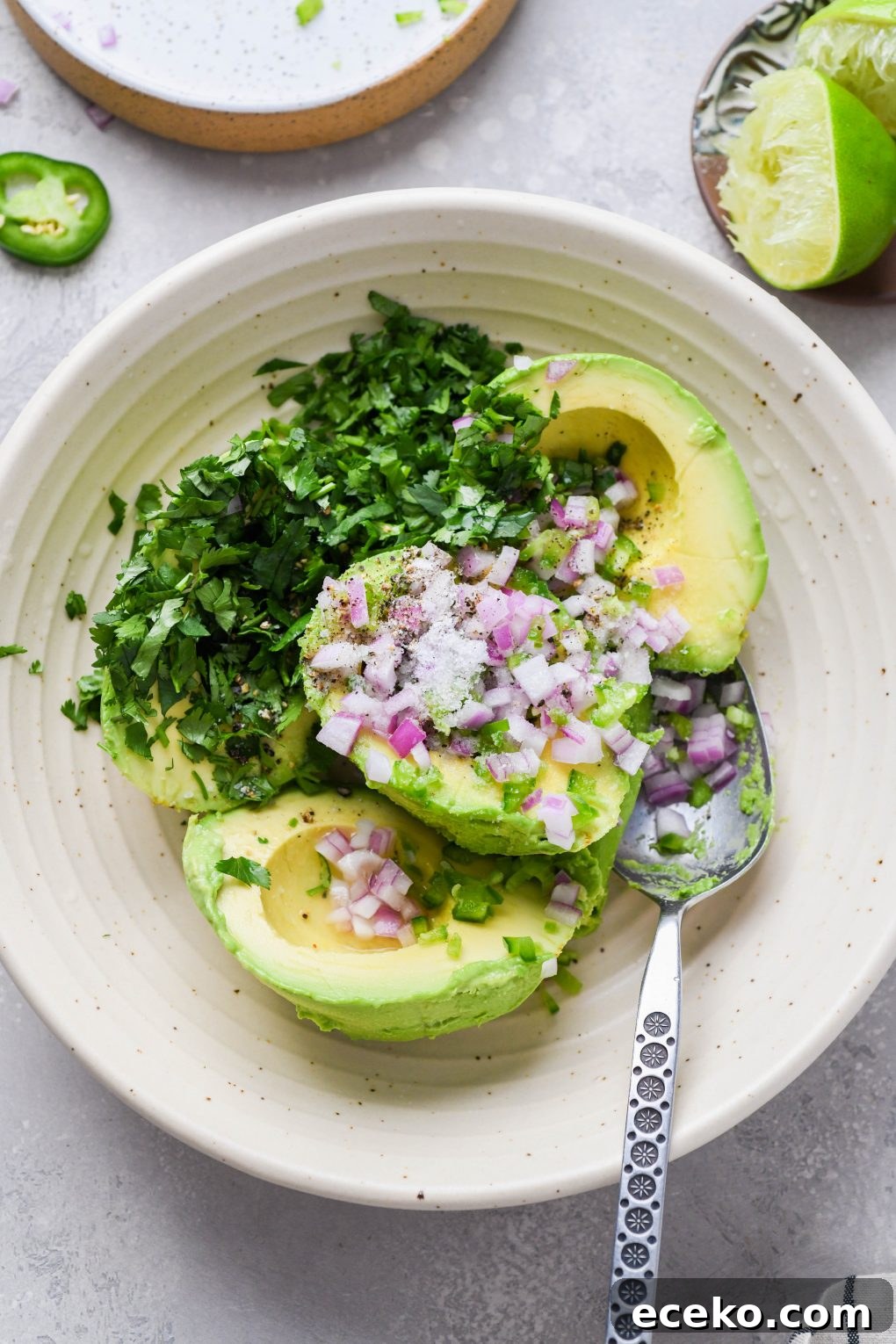Image of a large cream colored speckled bowl with ingredients for easy guacamole - avocado scooped out of the skin, diced red onion, jalapeno, cilantro, and salt and pepper. On a light grey background. 