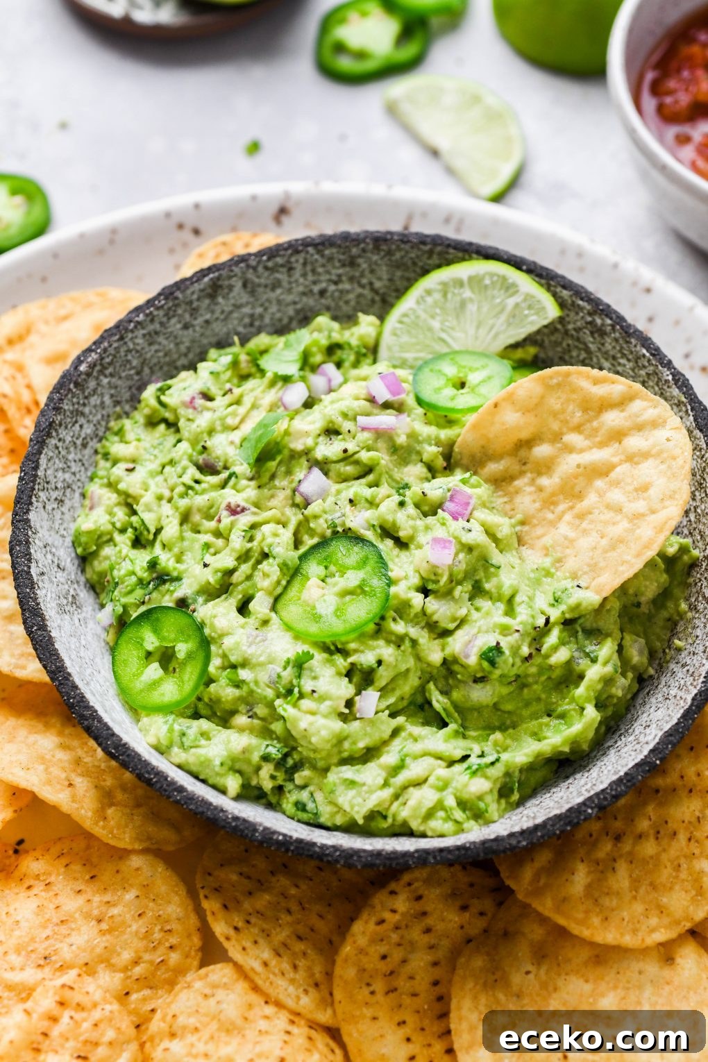Overhead shot of a small bowl of bright green guacamole. Guacamole is topped with some finely diced onion, thinly sliced jalapeño, and cilantro leaves, surrounded by round yellow tortilla chips. 