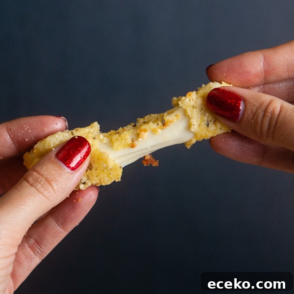 Low carb mozzarella stick being stretched apart by two female hands, showing a gooey cheese pull on black