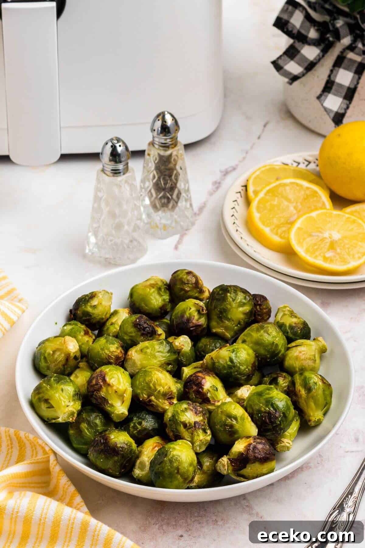 Green and juicy brussels sprouts in a white bowl in front of an air fryer with lemon slices on a plate. 