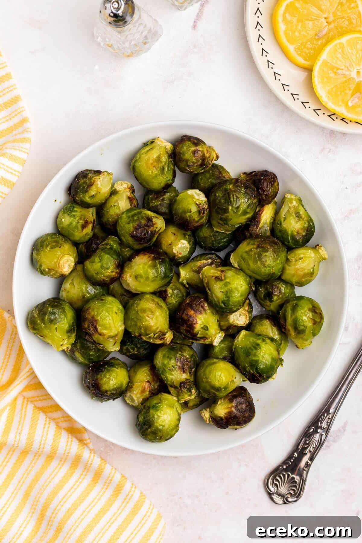 Green and juicy brussels sprouts in a white bowl in front of an air fryer with lemon slices on a plate.