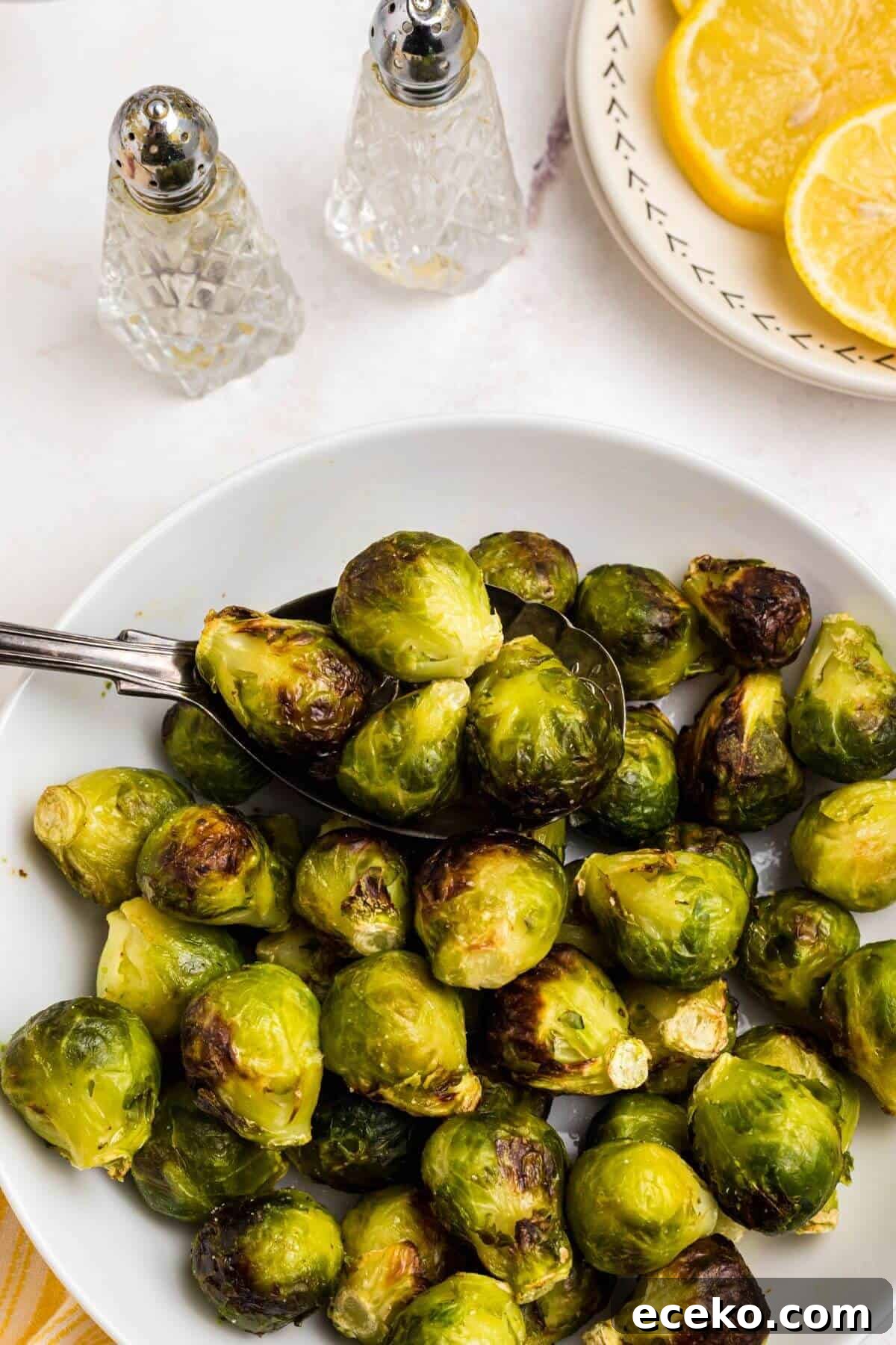 Green and juicy brussels sprouts in a white bowl in front of an air fryer with lemon slices on a plate and a spoon scooping out brussels sprouts. 