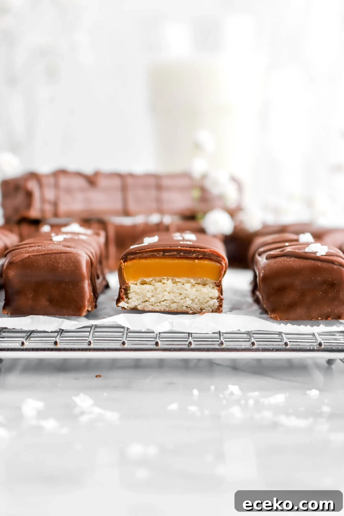 Homemade Twix Bars Recipe: Crispy Caramel Chocolate Cookies 4 close up of cut twix bar on wire cooling rack with more bars behind, a glass of milk, and white flowers