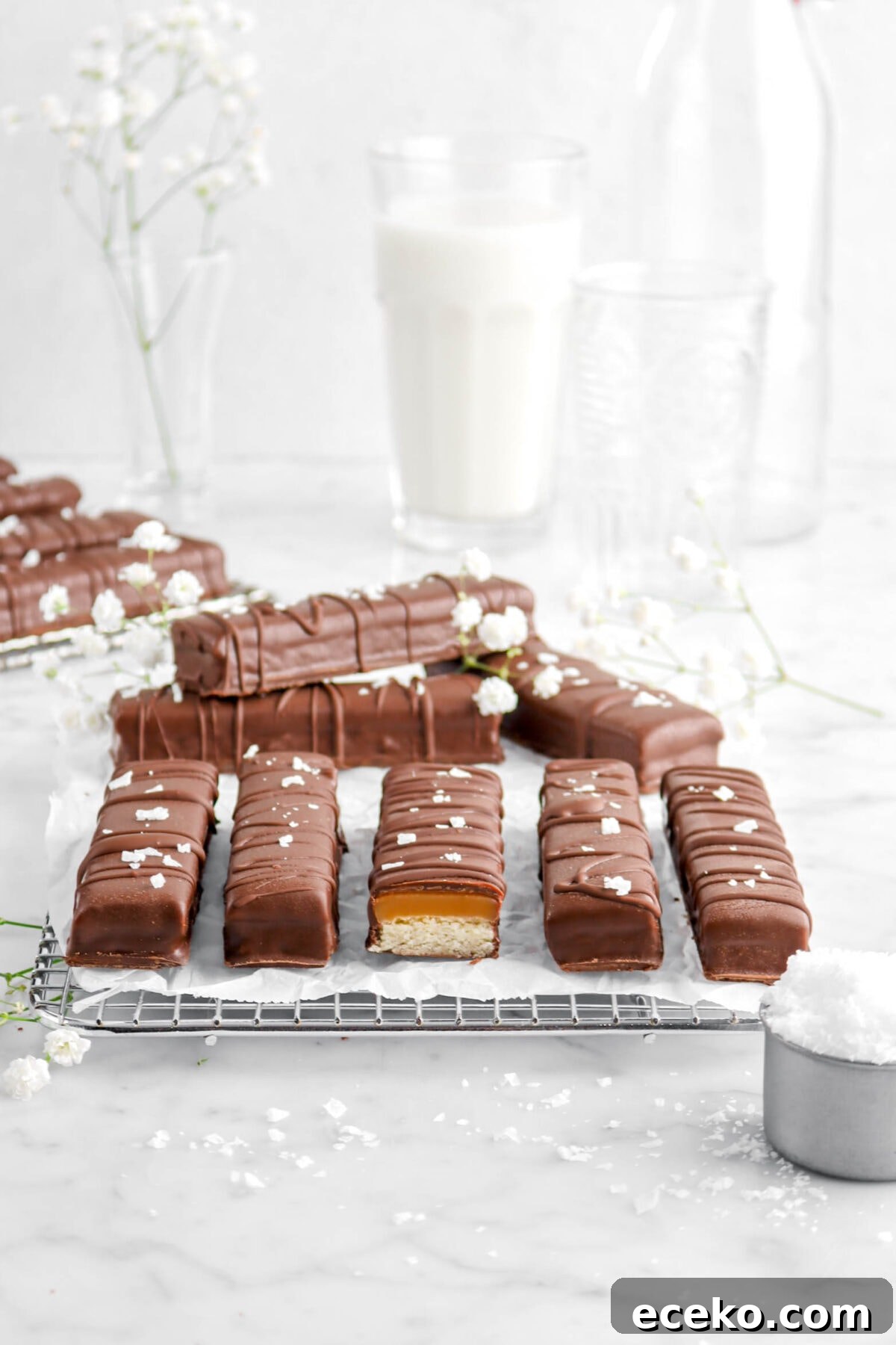 Homemade Twix Bars Recipe: Crispy Caramel Chocolate Cookies 5 pulled back shot of twix bars with one cut open on wire cooling rack with flowers, glass of milk, and flaked salt