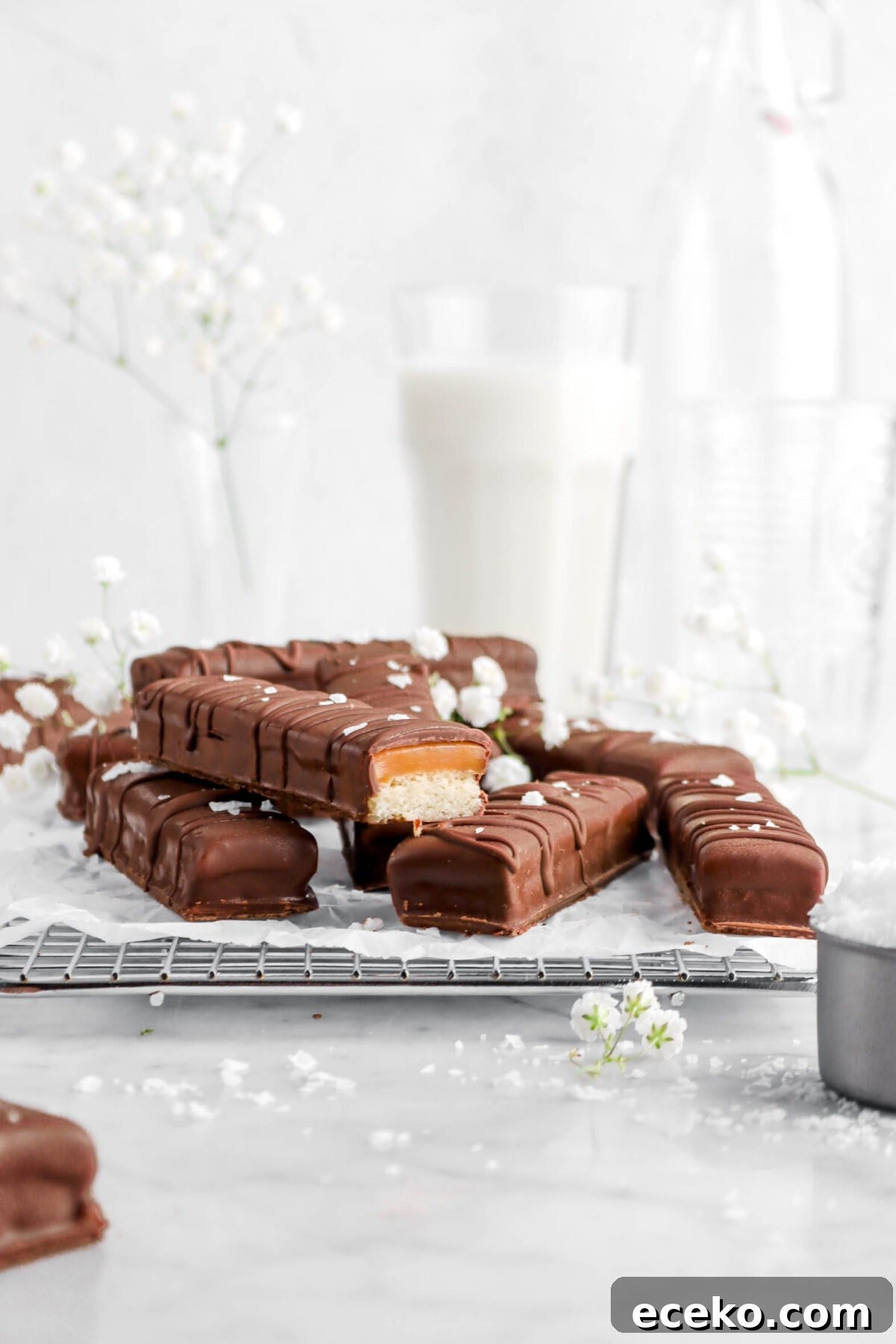 Homemade Twix Bars Recipe: Crispy Caramel Chocolate Cookies 7 homemade twix bars on wire cooling rack on parchment paper with one cut open, flaked salt, flowers, and a glass of milk behind