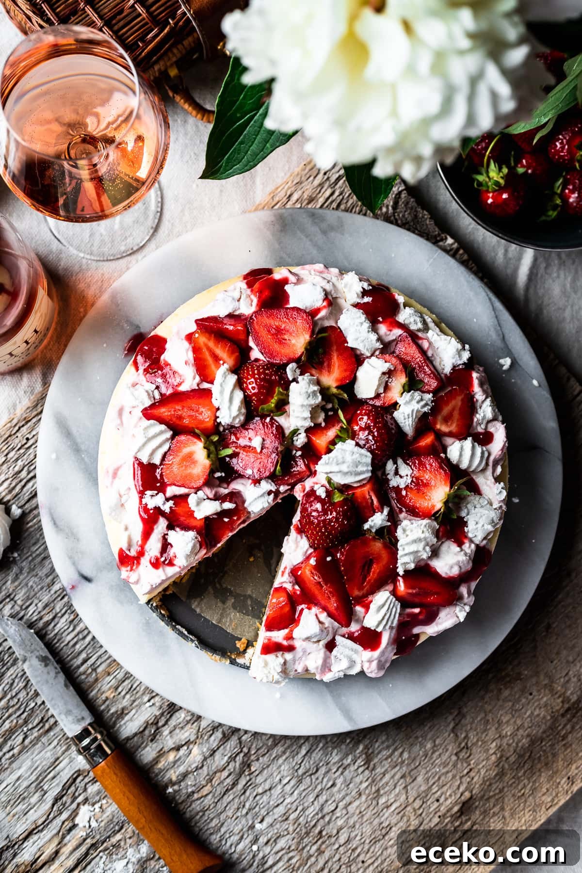 Top view of strawberry topped cheesecake with a slice missing. A rustic wooden board serves as a background. A glass of rosé wine sits nearby.