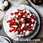 A top view of a vanilla cheesecake topped with whipped cream, strawberry coulis, fresh strawberries and rose water meringues on a marble platter and a rustic wooden cutting board.