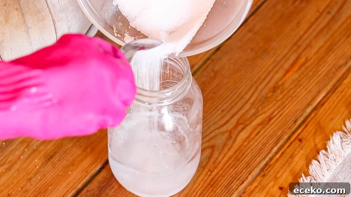 A gloved hand spooning lye into a mason jar filled with water.