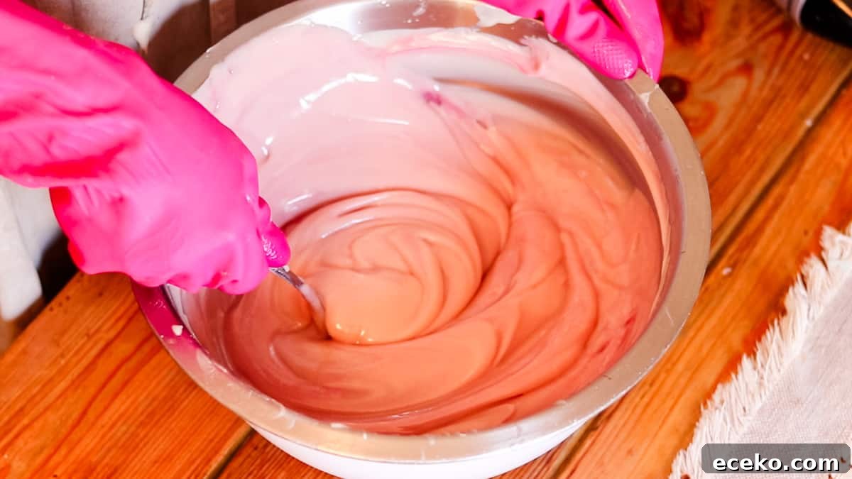 A woman wearing rubber gloves using a metal spoon to stir pink homemade soap batter in a large metal bow.
