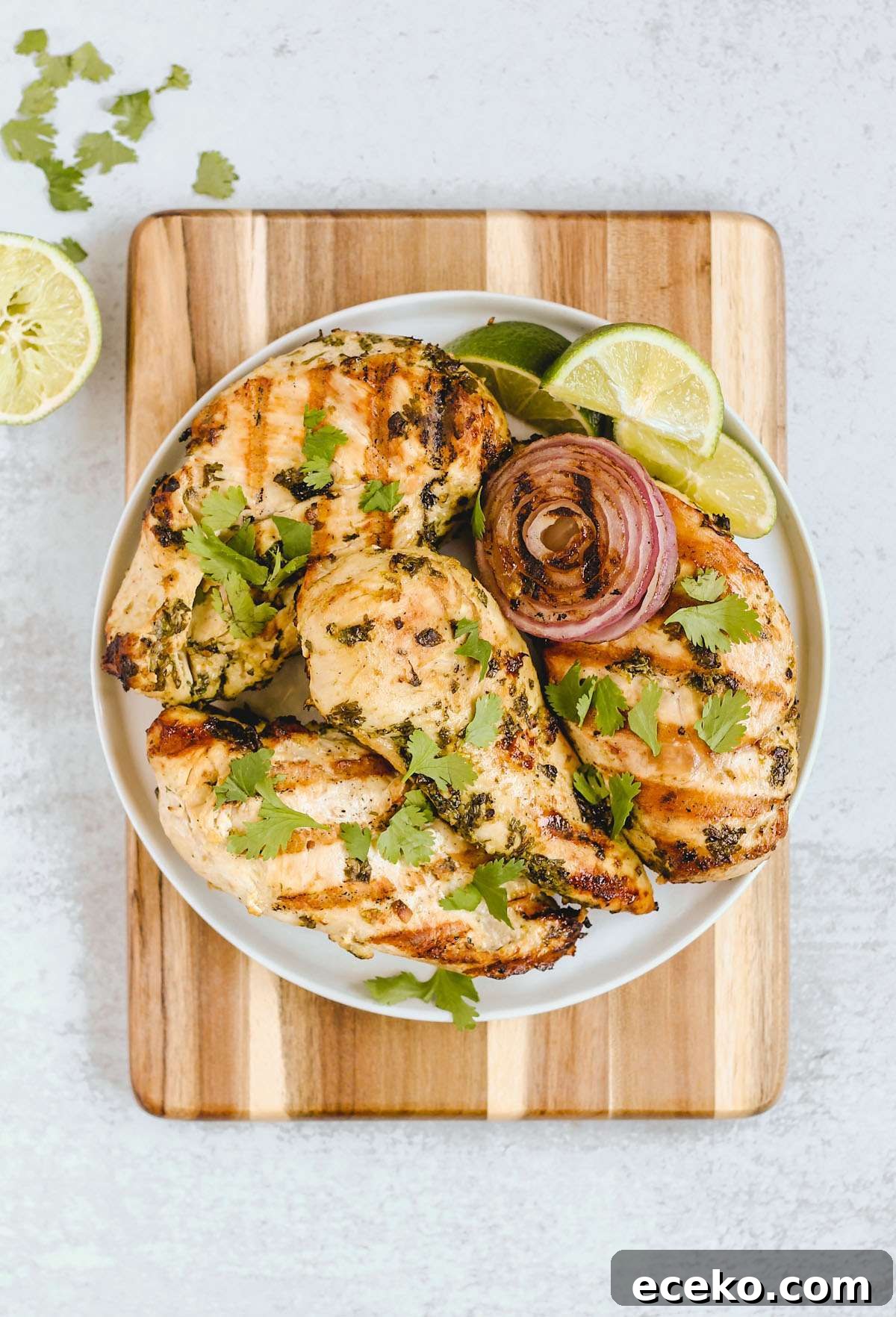 Four grilled cilantro lime chicken breasts with grilled onions and lime slices on a round plate on top of a wood cutting board.