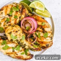 Four grilled cilantro lime chicken breasts with grilled onions and lime slices on a round plate.