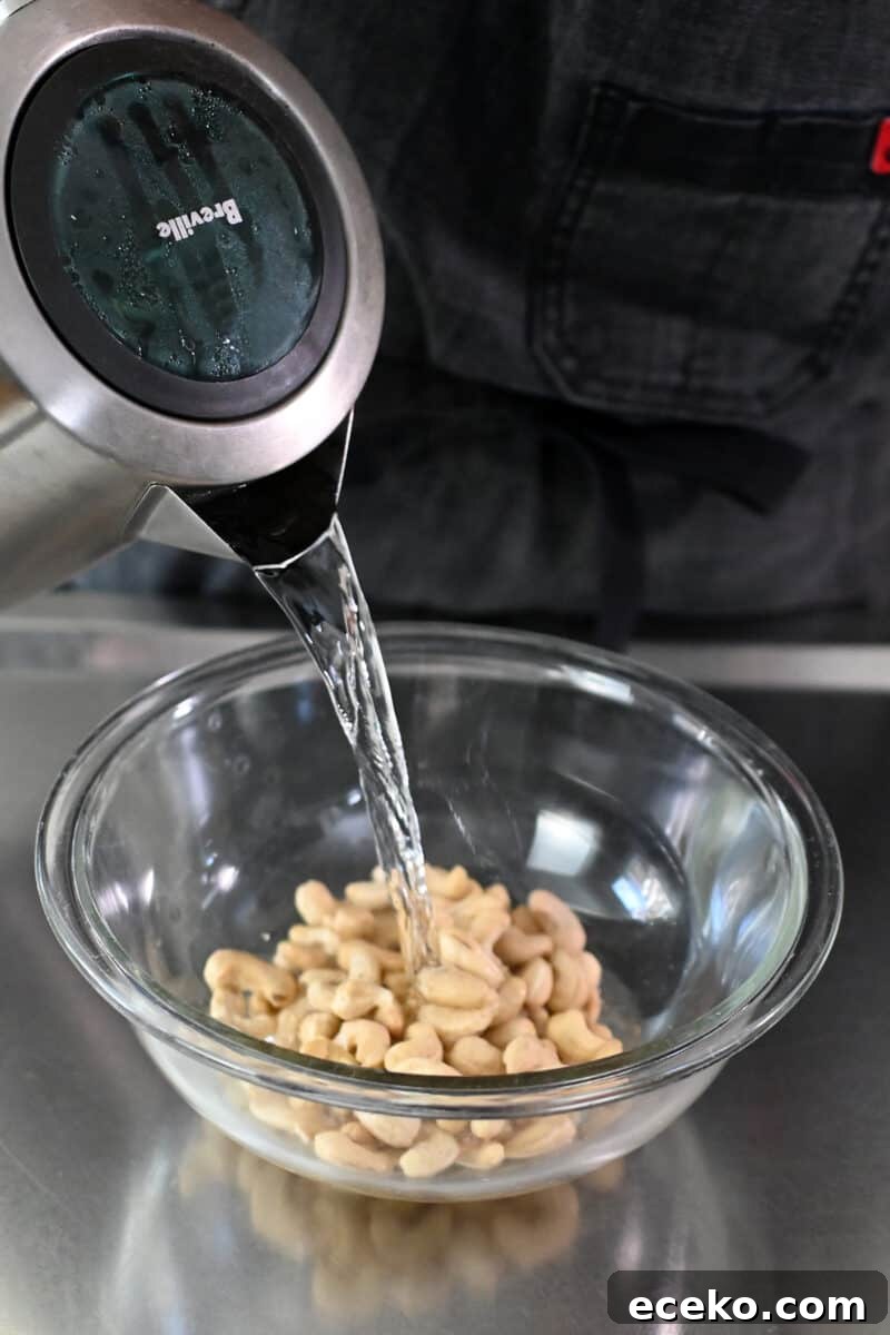 Hot water from a kettle is being poured on top of raw cashews in a clear glass bowl.