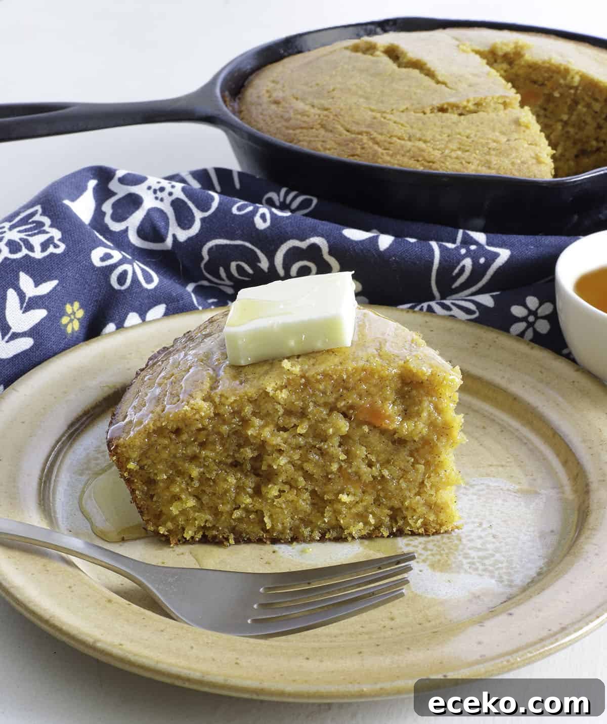 Slice of sweet potato cornbread with a pat of butter and a drizzle of honey on top.  Cast iron skillet in background.  Small bowl of honey on the side.