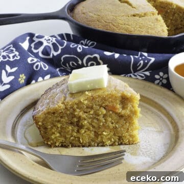 Slice of sweet potato cornbread with a pat of butter and a drizzle of honey on top. Cast iron skillet in background. Small bowl of honey on the side.