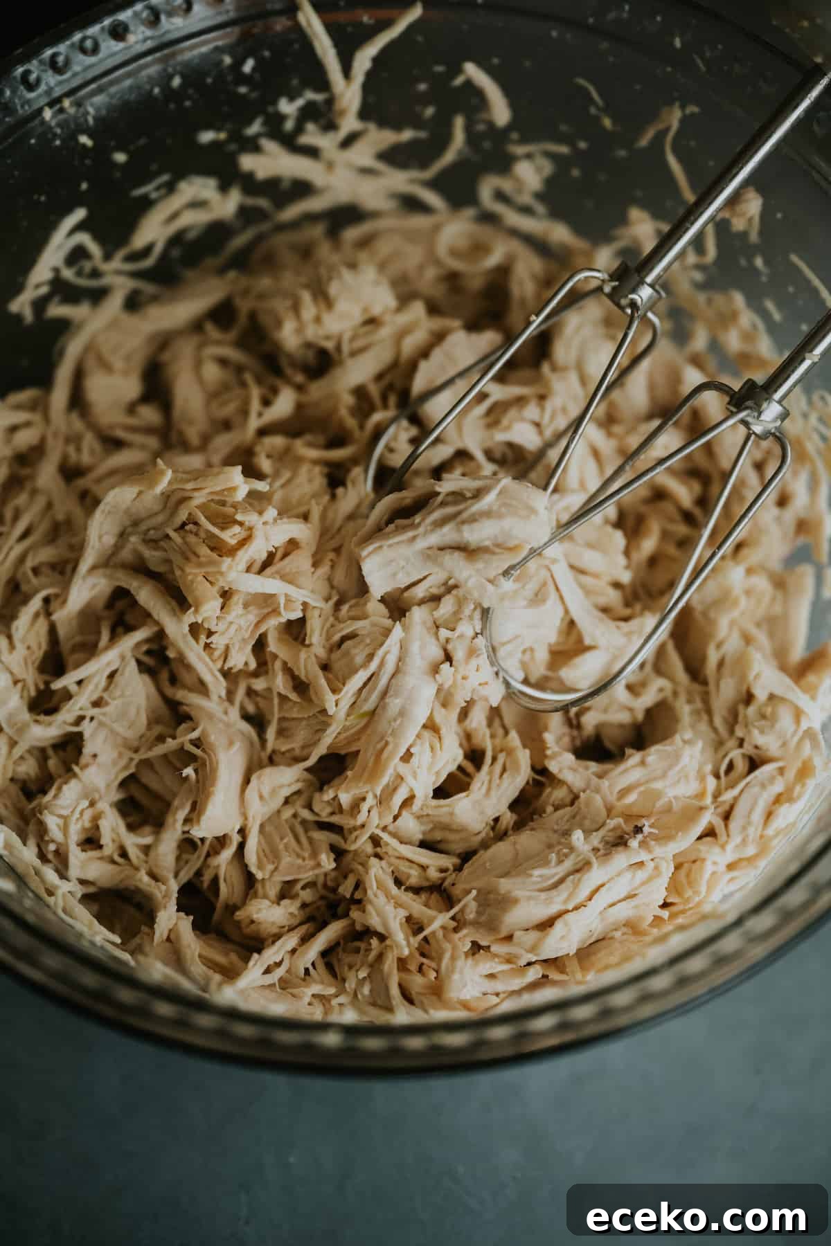 closeup on a mixing bowl of shredded chicken with an electric hand mixer.