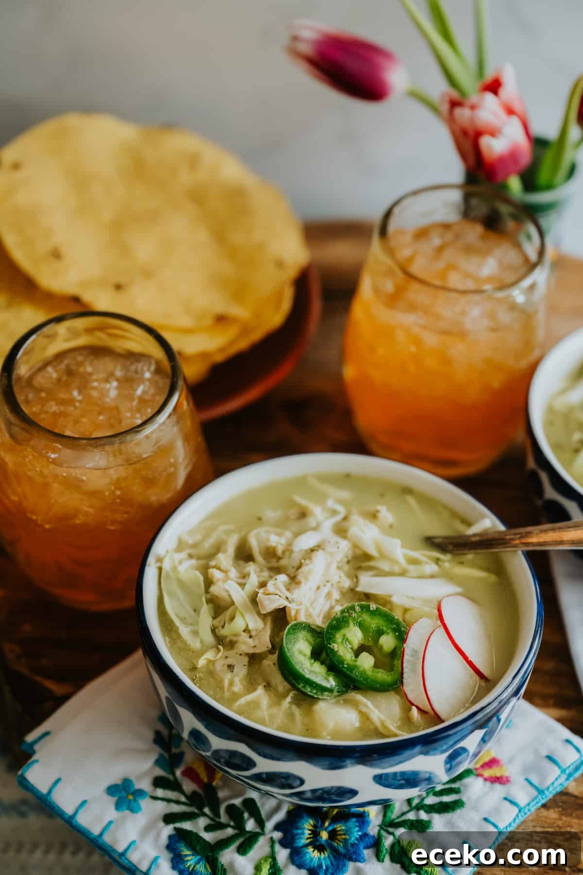 45 degree angle closeup shot of a bowl of chicken posole verde next to a glass of iced tea and a plate of tostadas.
