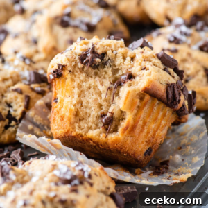 A close-up of a chocolate chip muffin with a bite taken out, showing its moist, fluffy texture and melty chocolate chunks—perfect for bakery muffins lovers—with more muffins and chocolate pieces in the background.